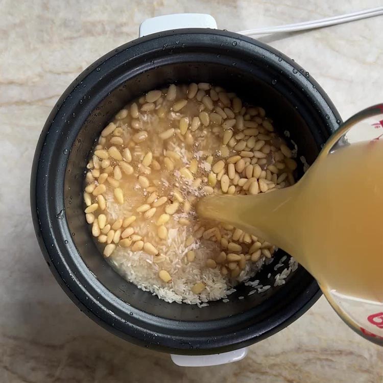 Pouring golden bone broth over rice and pine nuts in black rice cooker
