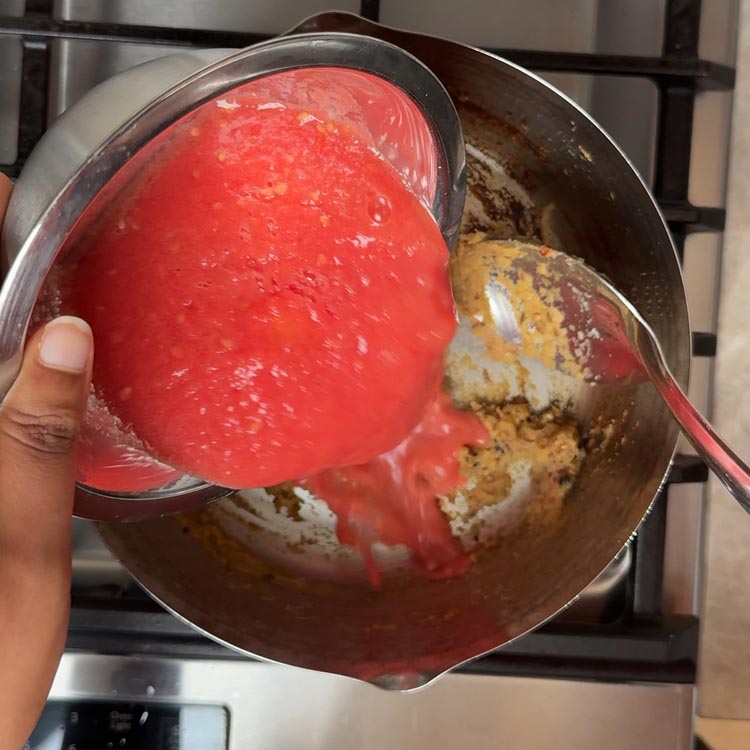Vibrant red tomato puree streaming from bowl into golden brown onion and spice mixture