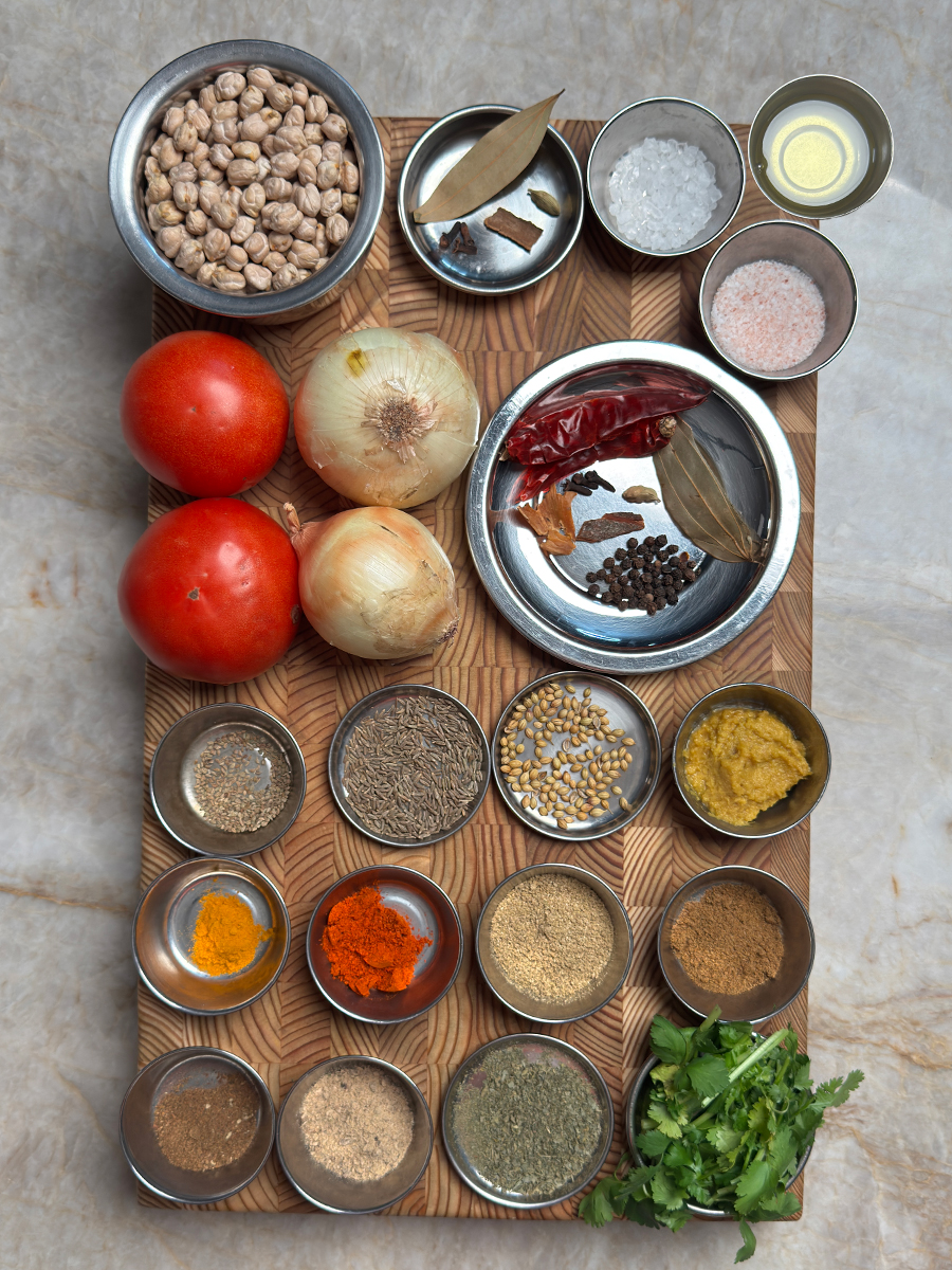 Dried chickpeas, fresh tomatoes, onions, whole spices, ground spices, and fresh coriander arranged on wooden board