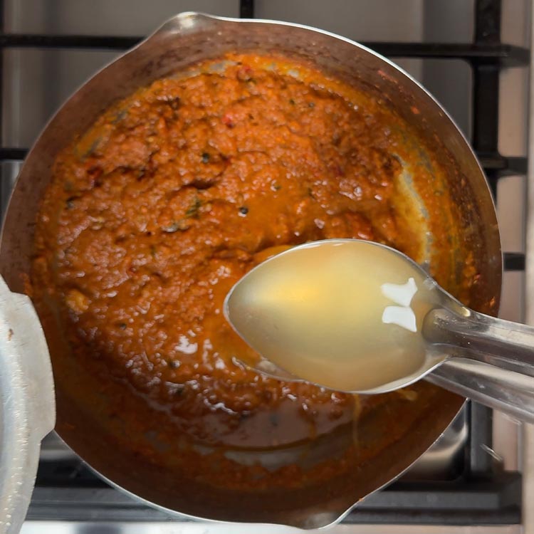 Ladle pouring pale amber chickpea cooking liquid into thick spiced tomato curry