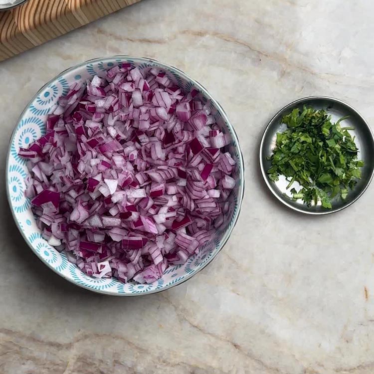 Finely chopped purple onions in decorative bowl next to fresh green coriander leaves in small metal dish