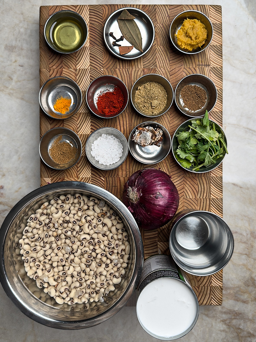 Measured ingredients for the curry laid out in small bowls on a wooden surface, showing turmeric, coriander powder, coconut milk can, and soaked black eyed peas