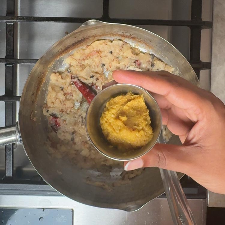 Small bowl of yellow ginger garlic paste being added to bubbling onion mixture with visible spices