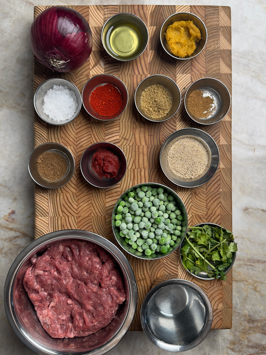 Organized ingredients on wooden board: ground lamb, spices, peas, onion, oil, and fresh herbs