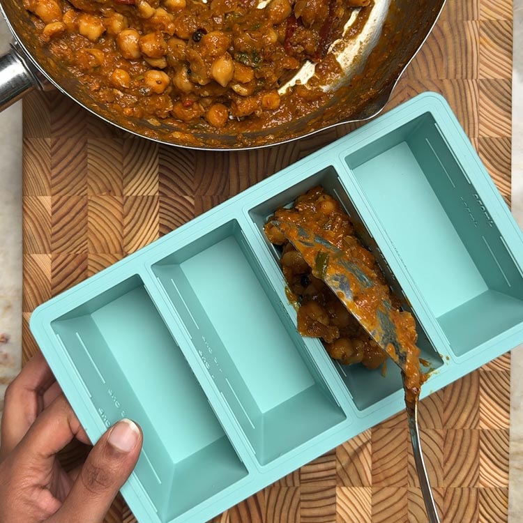Spooning chickpea curry from pan into turquoise silicone freezer compartments for meal prep