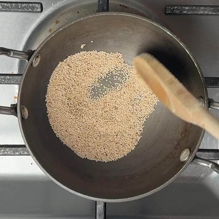 Golden poppy seeds being toasted in stainless steel pan with wooden spoon stirring