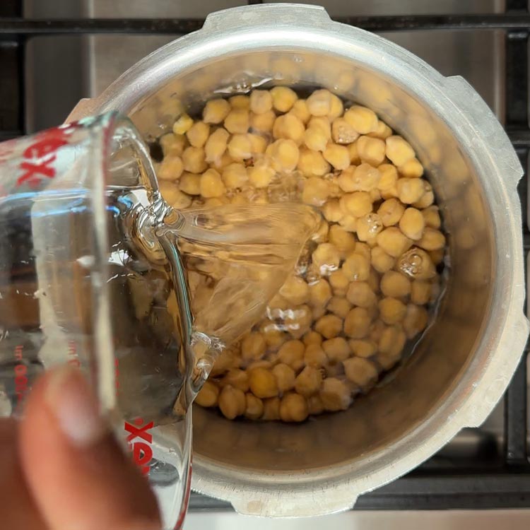 Hand pouring clear cooking liquid from measuring cup into pressure cooker with cooked chickpeas