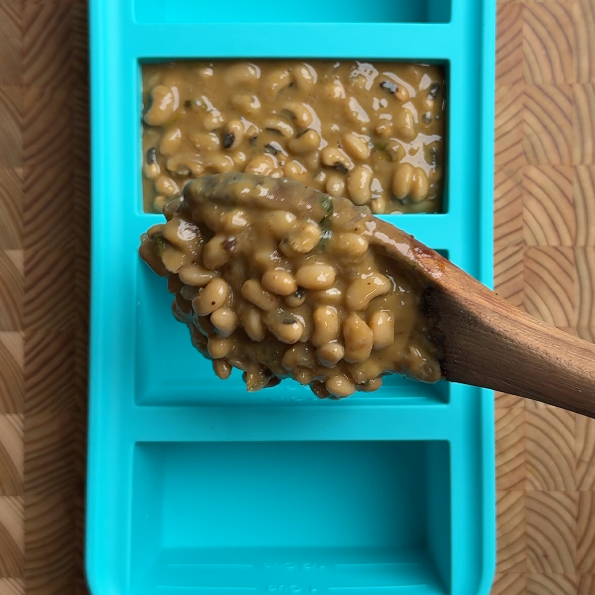 Wooden spoon lifting a portion of creamy black eyed peas curry from a frozen batch in a turquoise container