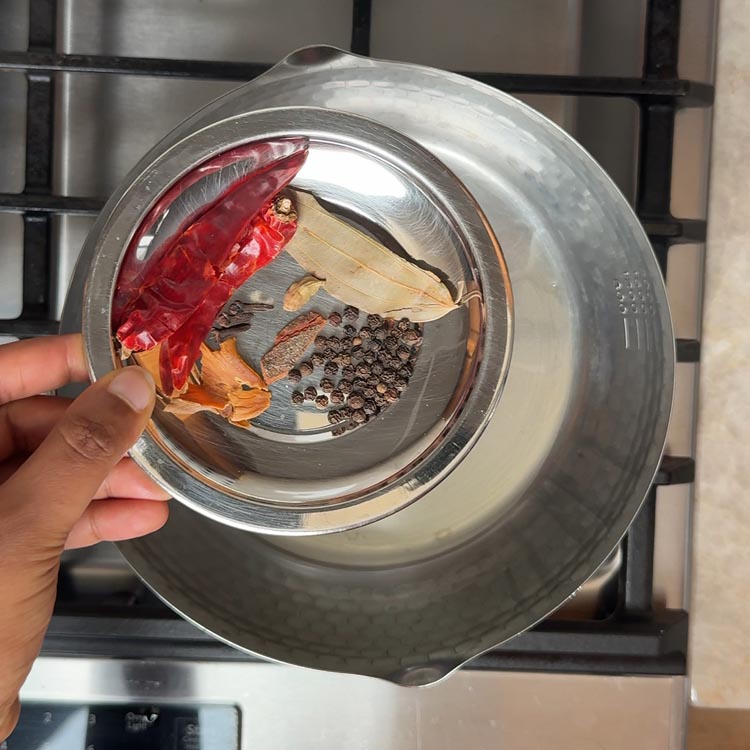 Red chilies, cinnamon stick, black peppercorns, and bay leaf sizzling in hot oil in steel pan