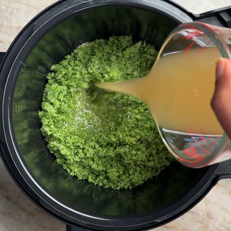 Pouring golden chicken bone broth from measuring cup into pressure cooker filled with riced broccoli