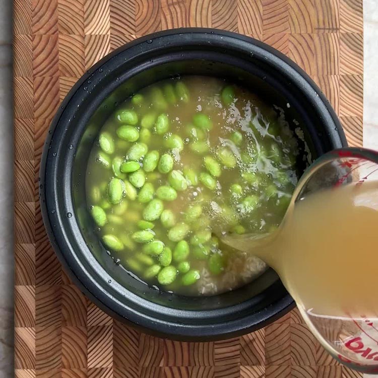 Chicken broth being poured into rice cooker with rice and edamame, combining all ingredients before cooking