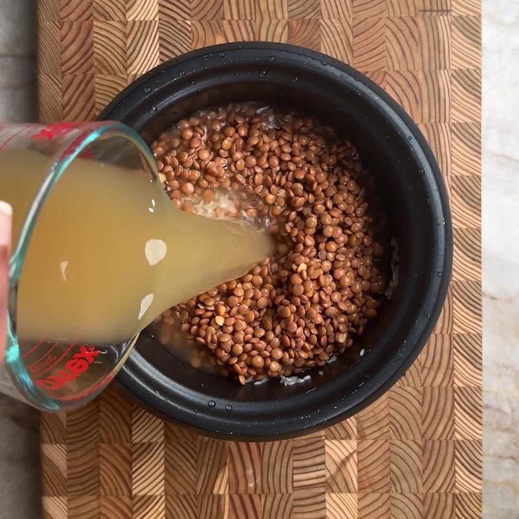 Pouring amber colored chicken bone broth into pot with soaked brown lentils