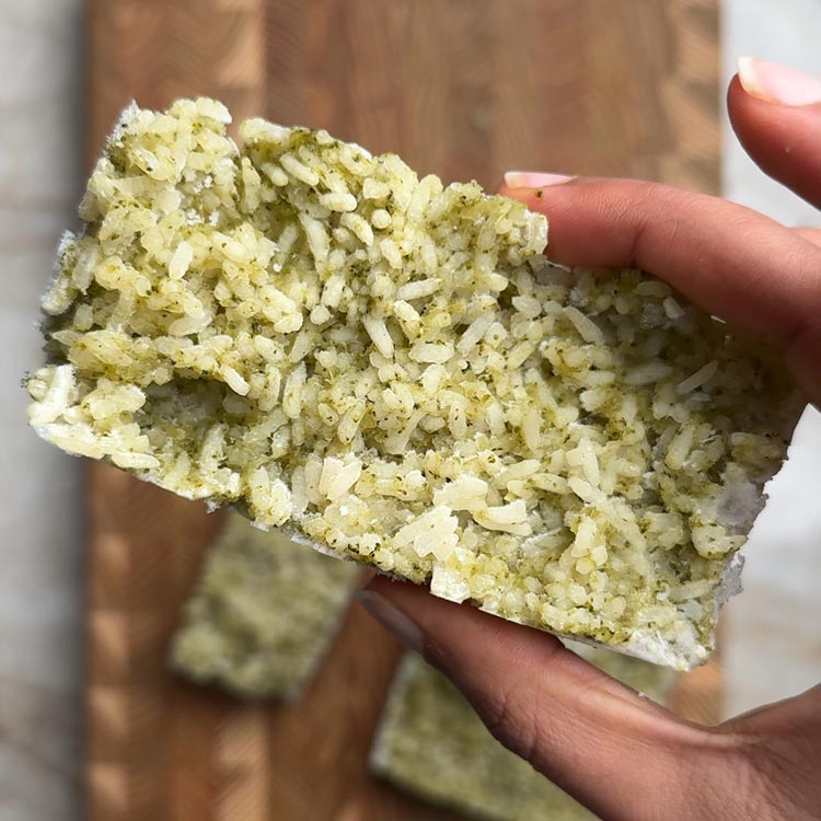 Hand holding single frozen rice cube showing green broccoli flecks throughout white rice