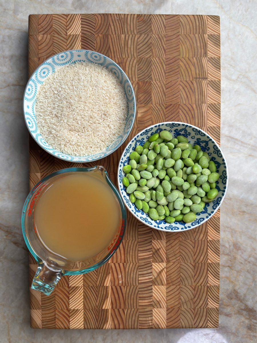 Three ingredients arranged for rice cooker recipe: white rice, frozen edamame, and chicken broth in separate bowls on a wooden cutting board