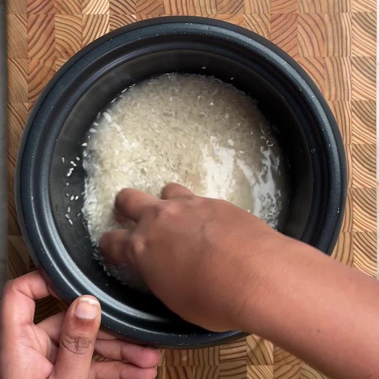 Hand rinsing uncooked white rice in a rice cooker bowl, removing excess starch until water runs clear