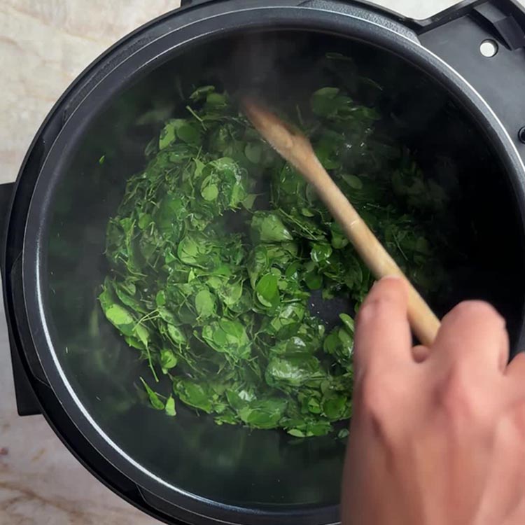 Fresh moringa leaves wilting in black Instant Pot inner pot while being stirred with wooden spoon