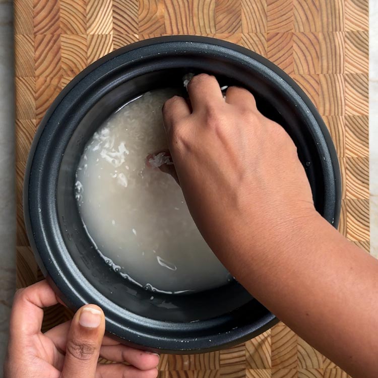 Hands rinsing white rice grains in cloudy water inside pressure cooker insert
