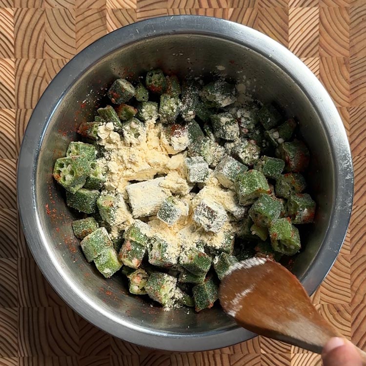 Besan chickpea flour sprinkled over seasoned okra in metal bowl with wooden spoon