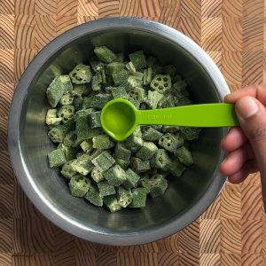 Hand holding green measuring spoon adding oil to frozen okra in metal mixing bowl