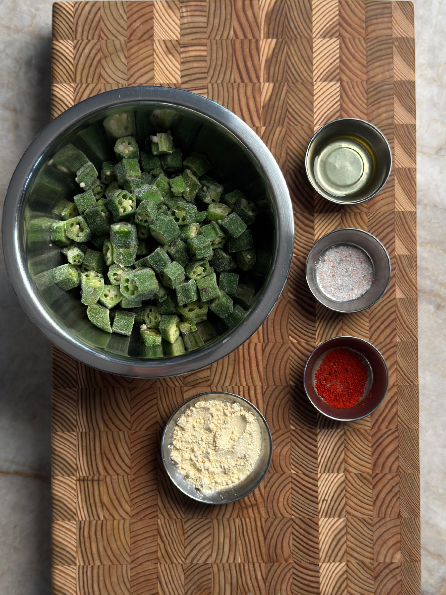 Frozen okra in metal bowl with besan powder, chili powder, salt and oil in small bowls on wooden cutting board