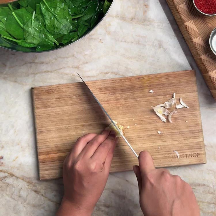 Hands mincing a garlic clove on a wooden cutting board with spinach soaking nearby