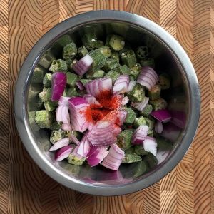 Frozen okra and red onion slices being tossed with oil and spices in mixing bowl
