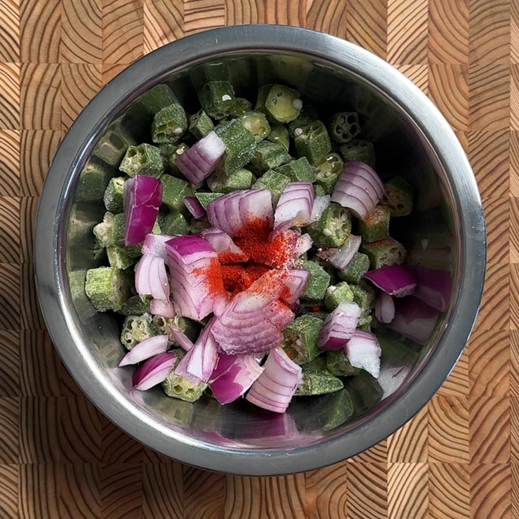 Frozen okra and red onion slices being tossed with oil and spices in mixing bowl