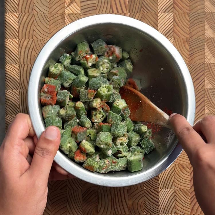 Hands mixing frozen okra with oil and chili powder using wooden spoon in metal bowl