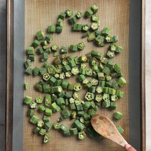 Green okra pieces spread on a silicone baking mat with a wooden spoon for spreading
