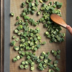 Frozen okra pieces spread in single layer on silicone mat lined sheet pan with wooden spoon