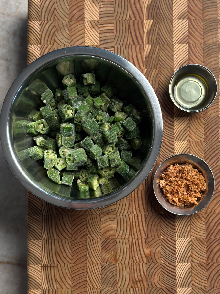 Frozen okra in metal bowl with garlic coconut podi powder and oil on wooden cutting board
