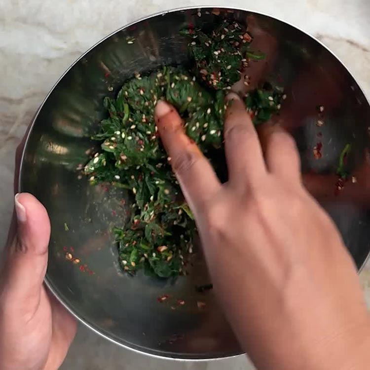 Hands massaging gochugaru, sesame seeds, and soy sauce into blanched spinach