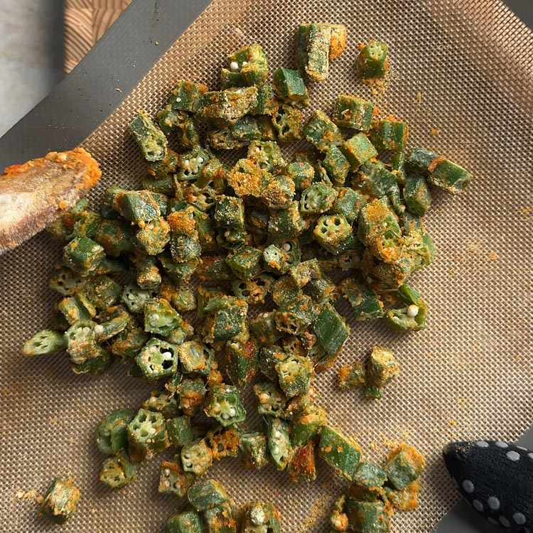 Wooden spoon tossing baked besan okra on sheet pan to evenly distribute besan coating