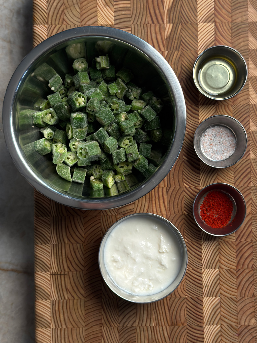 Frozen okra in a metal bowl with small dishes of yogurt, oil, salt, and red chili powder on a wooden board