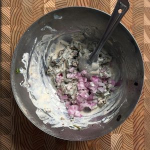 Yogurt coated okra in a metal bowl with diced pink onions and a black spatula