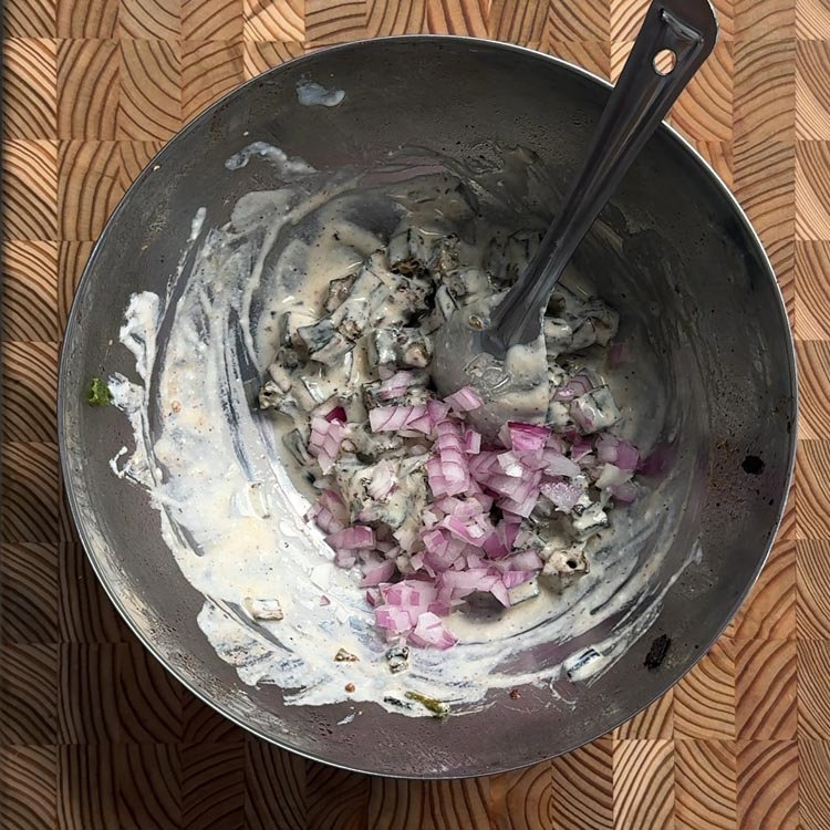 Yogurt coated okra in a metal bowl with diced pink onions and a black spatula