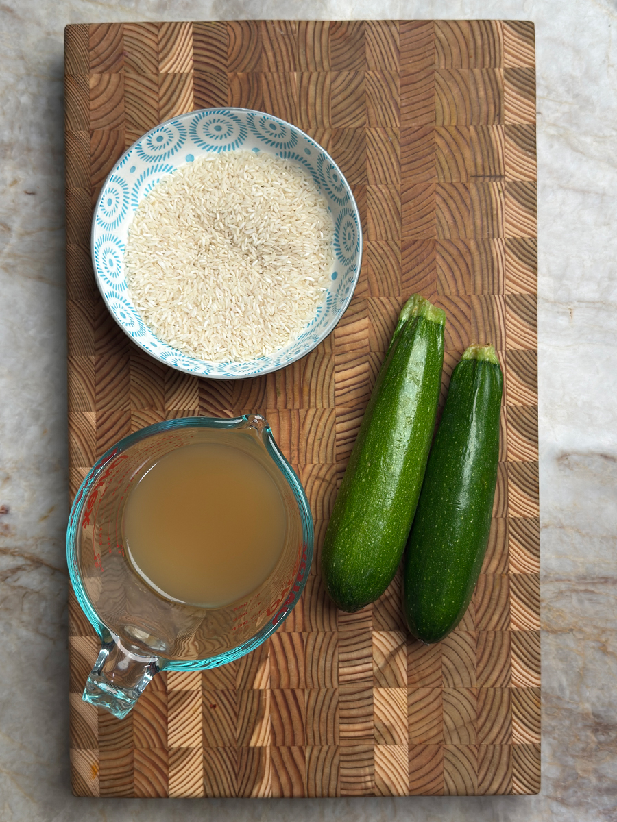 Bowl of white rice, measuring cup with bone broth, and two fresh zucchinis on a wooden cutting board