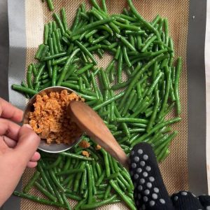 Sprinkling coconut garlic podi with a wooden spoon onto roasted green beans on a sheet pan