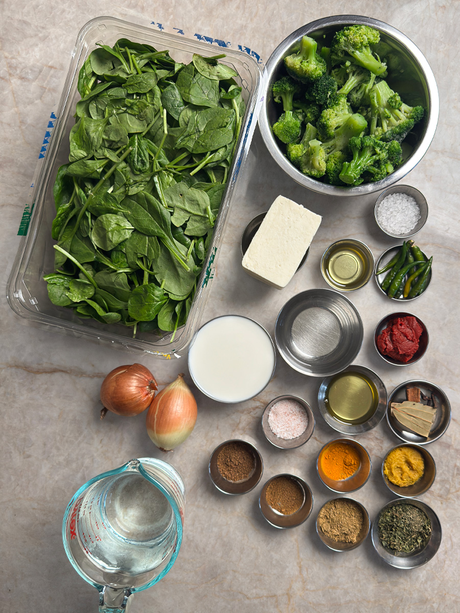 Fresh spinach in container, broccoli florets, paneer block, milk, onions, and spices arranged on counter