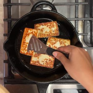 Four golden brown paneer strips frying in cast iron skillet with spatula flipping
