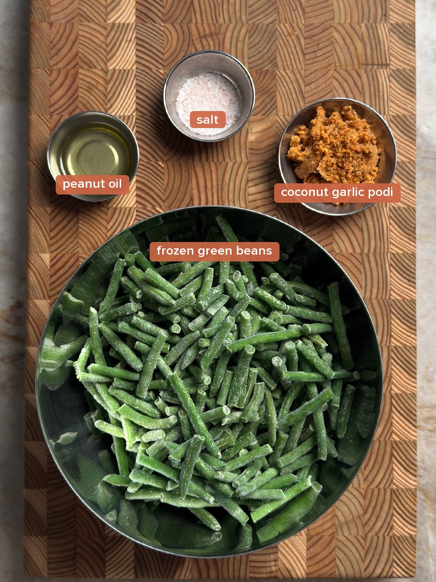 Frozen green beans in a metal bowl with small bowls of oil, pink salt, and coconut garlic podi on a wooden cutting board