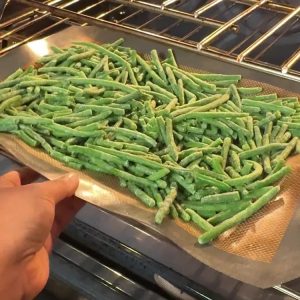 Hand sliding a sheet pan of seasoned green beans into a hot oven on a silicone baking mat
