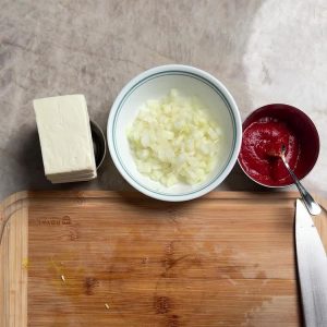 Paneer block, chopped onions in bowl, and tomato passata on wooden cutting board