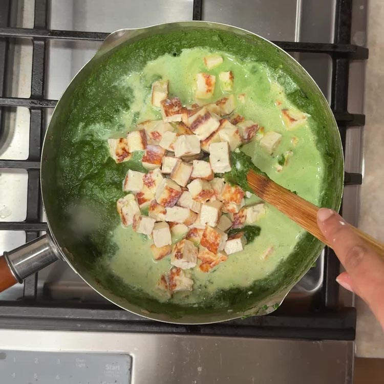 Golden paneer cubes added to bright green palak gravy in pan with wooden spoon