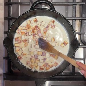 Cubed fried paneer pieces soaking in warm milk in cast iron skillet with wooden spoon