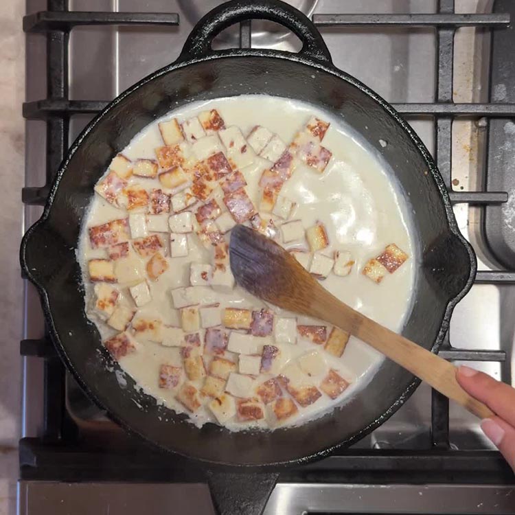 Cubed fried paneer pieces soaking in warm milk in cast iron skillet with wooden spoon