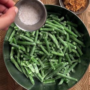 Hand sprinkling pink salt from a small bowl onto frozen green beans, coconut podi visible in corner