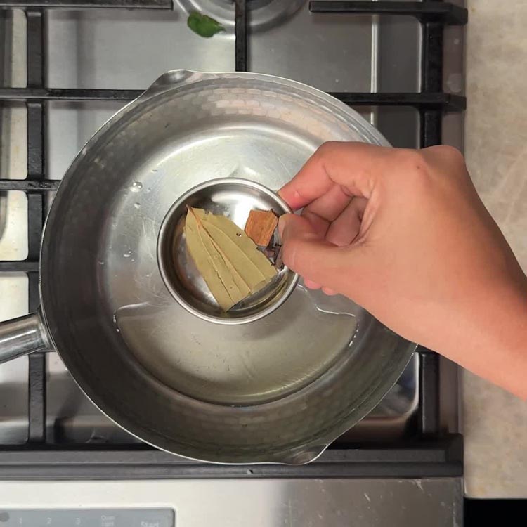 Bay leaf, cinnamon stick, and cloves being added to hot oil in stainless steel pan