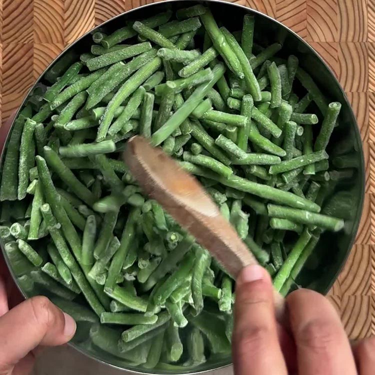 Hands tossing frozen green beans with a wooden spoon in a metal mixing bowl