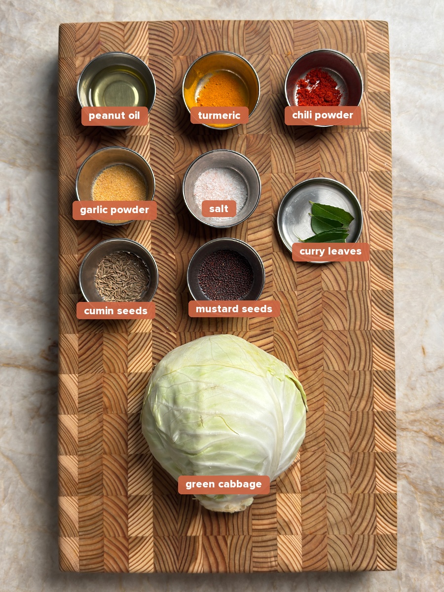 Whole green cabbage with small bowls of turmeric, chili powder, garlic powder, salt, cumin seeds, mustard seeds, curry leaves, and oil